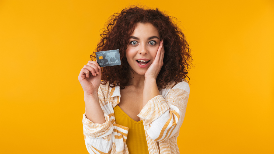 Excited woman holding a credit card and looking surprised against a bright yellow background.