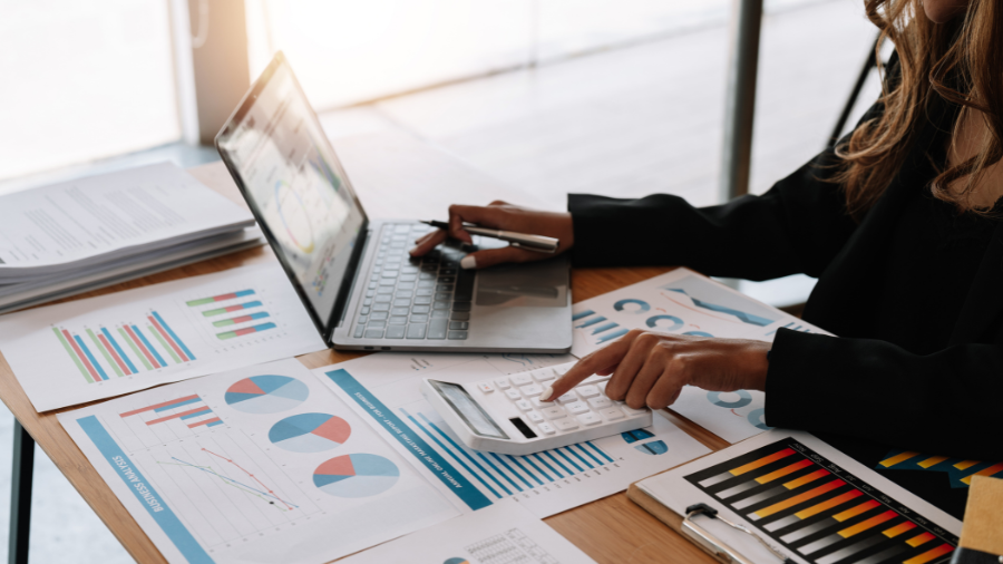 Person using a calculator and laptop surrounded by printed financial charts and graphs at a desk.