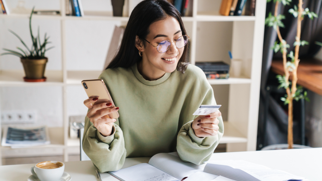 A woman wearing glasses and a green sweatshirt smiles while holding a phone and a credit card at a desk with an open notebook and coffee.